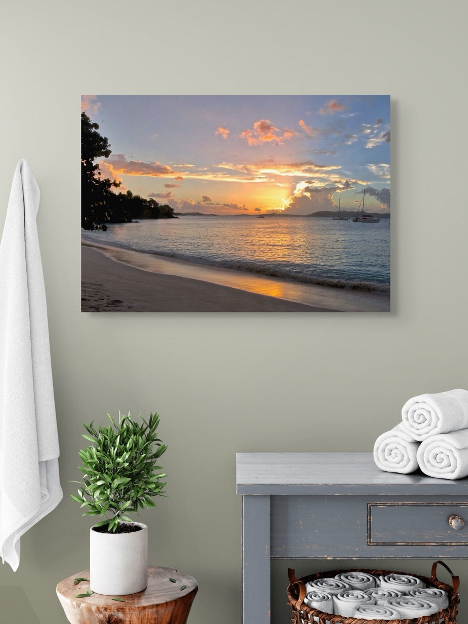A tropical sunset canvas print  on a wall above a dresser with towels and a plant.