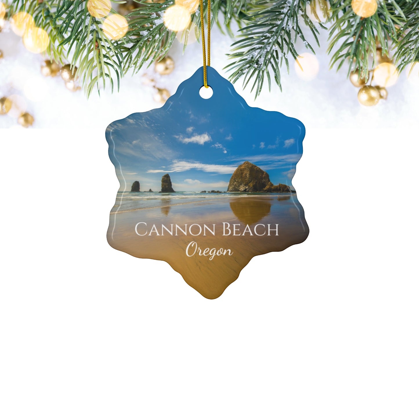 Ceramic holiday ornament featuring a photograph of Haystack Rock at Cannon Beach, Oregon, with blue sky reflections on the shoreline hanging among Christmas lights and tree branches.