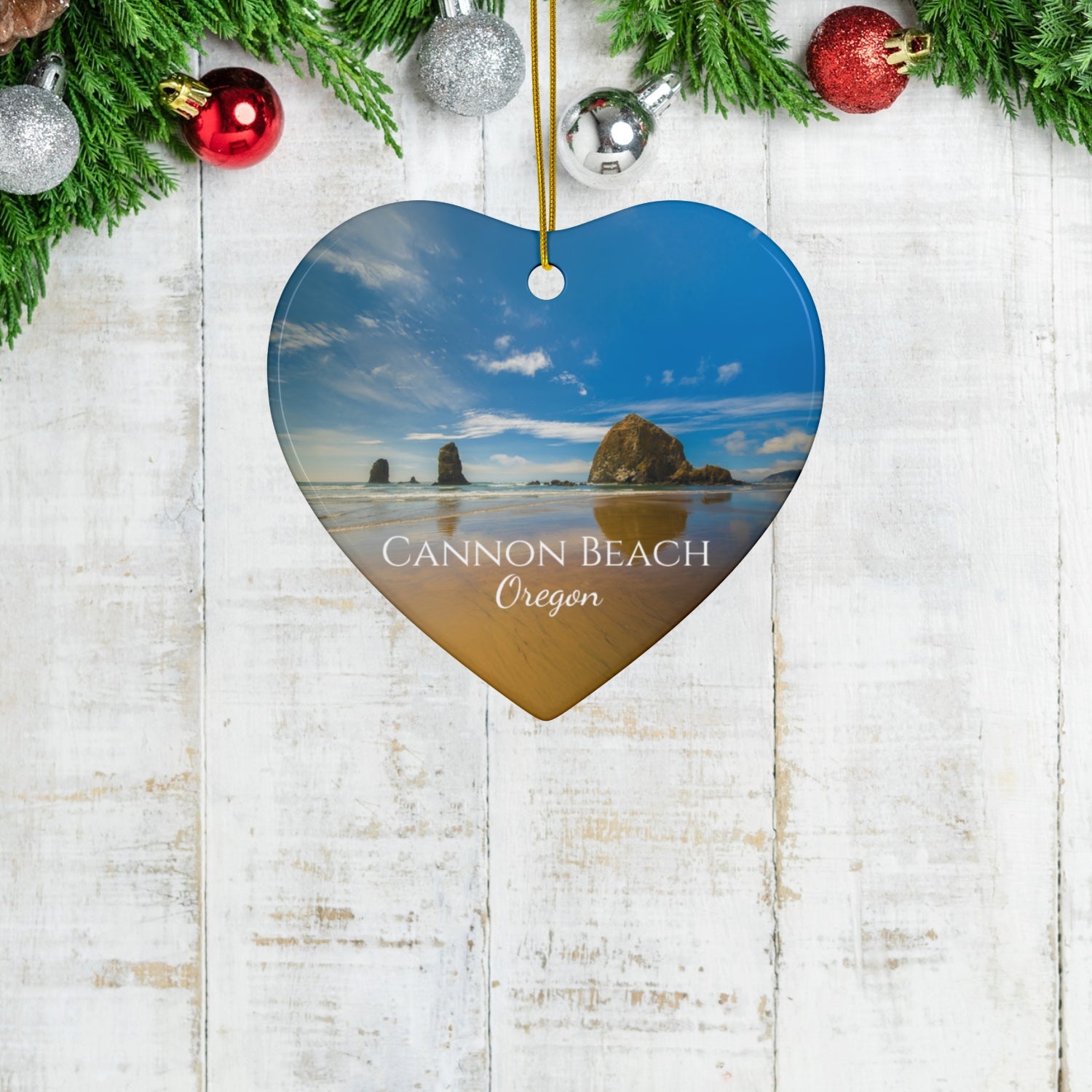 Heart-shaped ceramic holiday ornament featuring a photograph of Haystack Rock at Cannon Beach, Oregon, with blue sky reflections on the shoreline on a wooden surface with Christmas decorations.