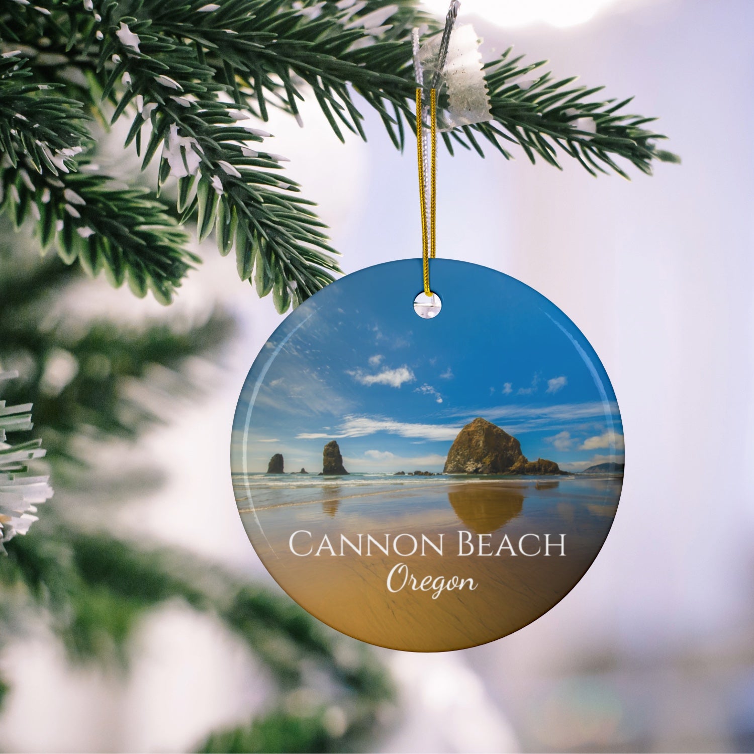 Round ceramic holiday ornament featuring a photograph of Haystack Rock at Cannon Beach, Oregon, with blue sky reflections on the shoreline hanging on a Christmas tree.
