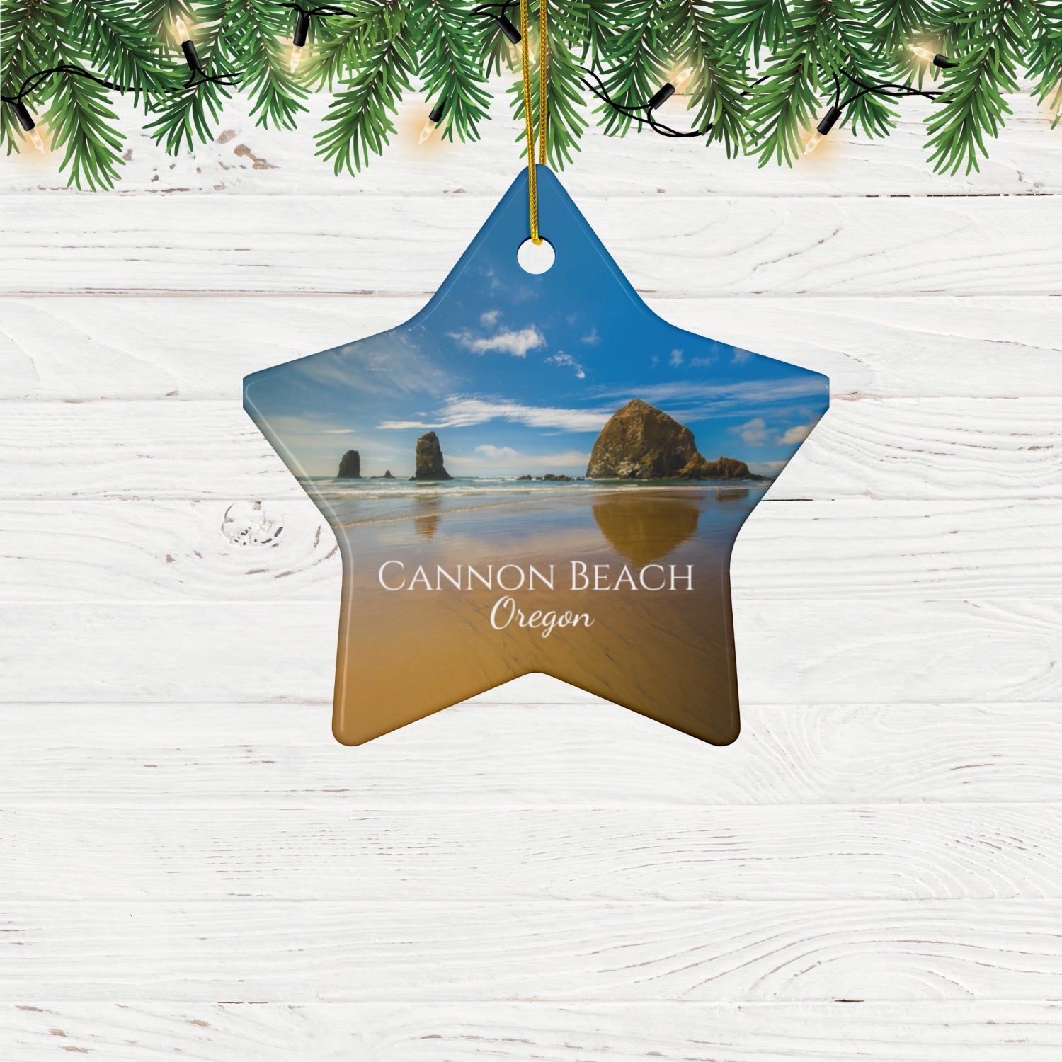 Star-shaped ceramic holiday ornament featuring a photograph of Haystack Rock at Cannon Beach, Oregon, with blue sky reflections on the shoreline on a wooden surface with Christmas tree branches.