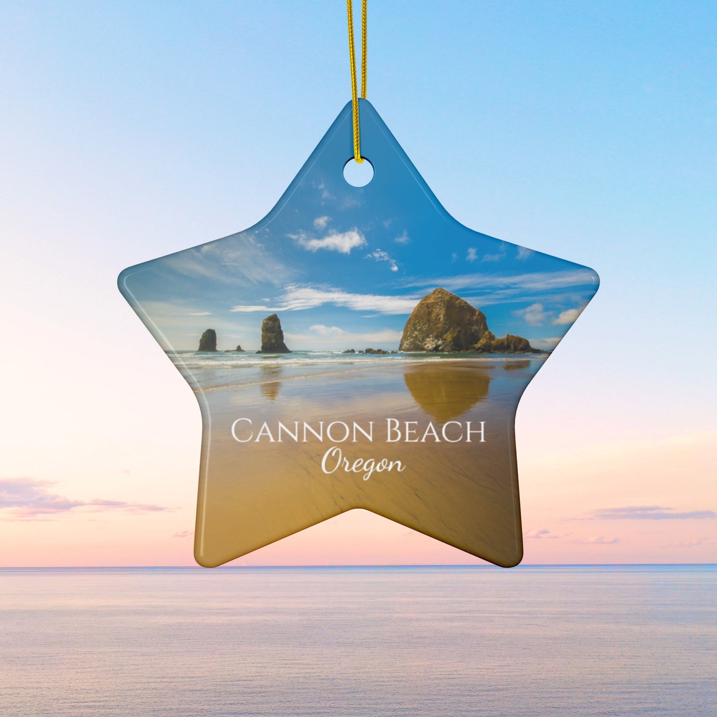 Star-shaped ceramic holiday ornament featuring a photograph of Haystack Rock at Cannon Beach, Oregon, with blue sky reflections on the shoreline