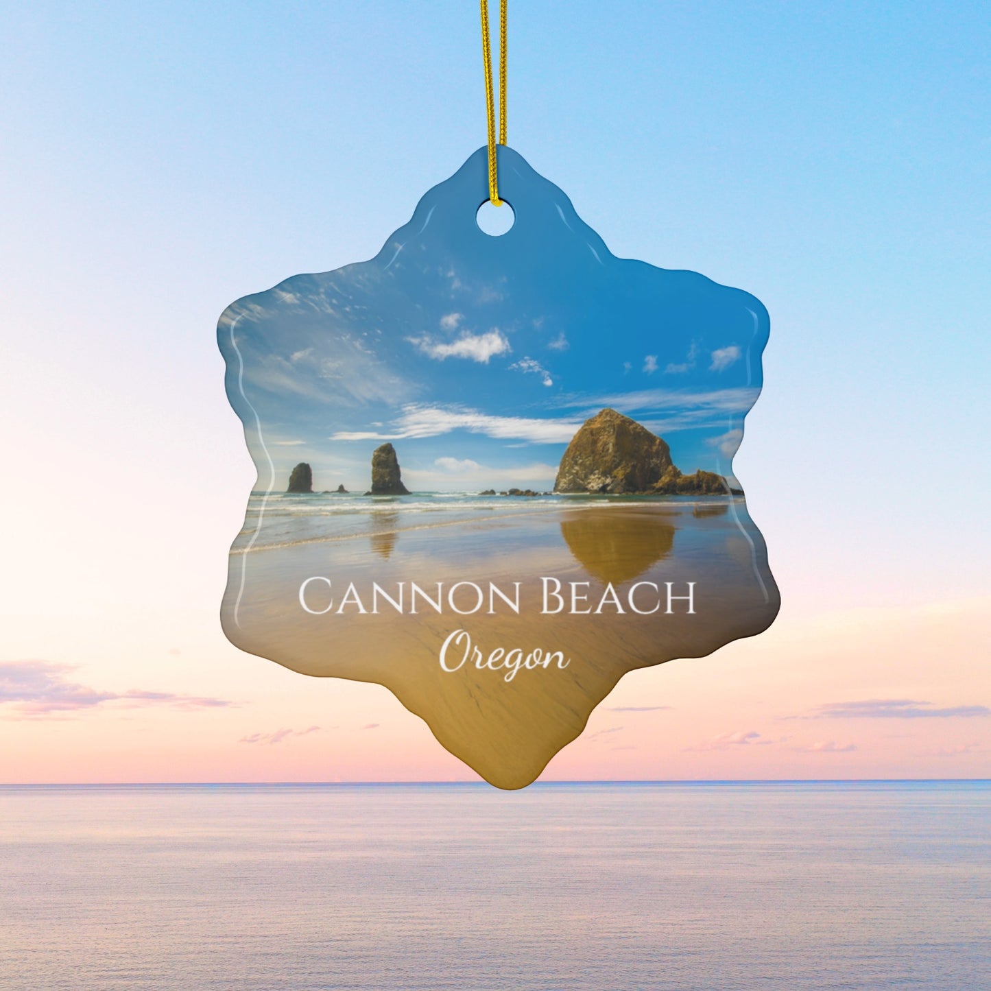 Ceramic holiday ornament featuring a photograph of Haystack Rock at Cannon Beach, Oregon, with blue sky reflections on the shoreline
