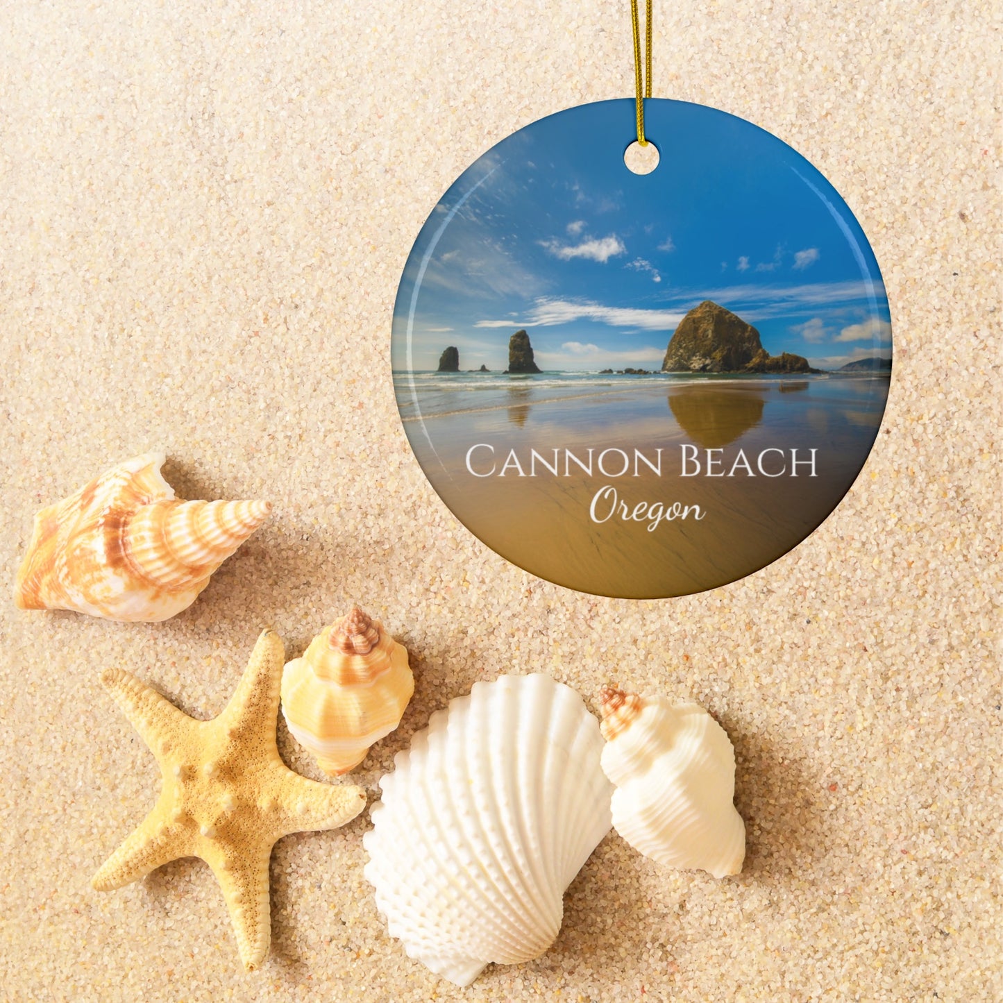 Round ceramic holiday ornament featuring a photograph of Haystack Rock at Cannon Beach, Oregon, with blue sky reflections on the shoreline, on sand with shells and a starfish.