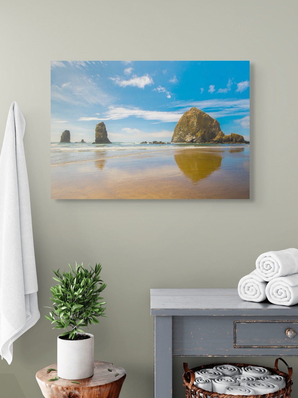 A canvas print featuring a view of Haystack Rock and other rock off the coast of Cannon Beach, Oregon on a wall above a dresser with towels and a plant.