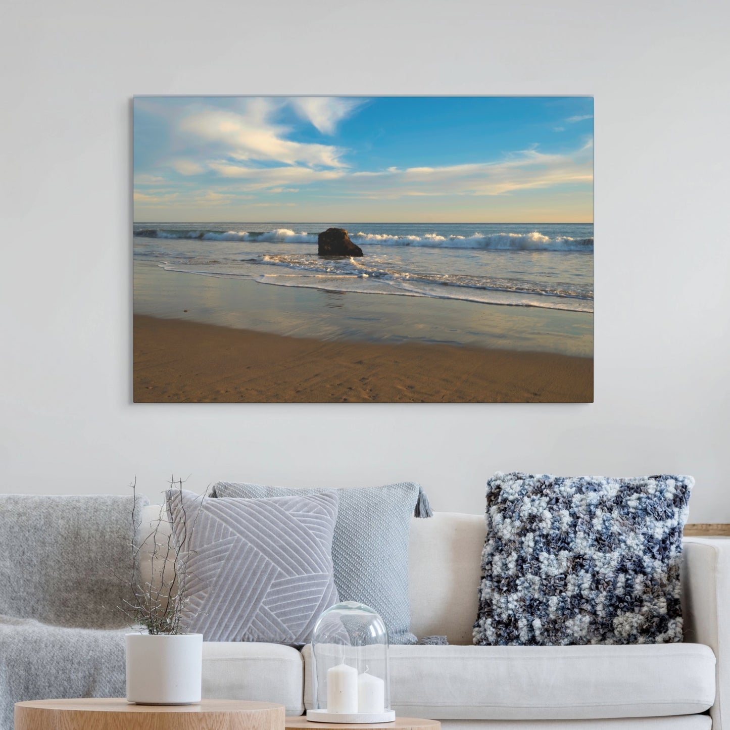 Solitary rock on a California beach with ocean wave foam and sky reflection canvas print on a wall above a sofa with decorative pillows.