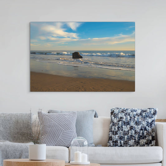 Solitary rock on a California beach with ocean wave foam and sky reflection canvas print on a wall above a sofa with decorative pillows.