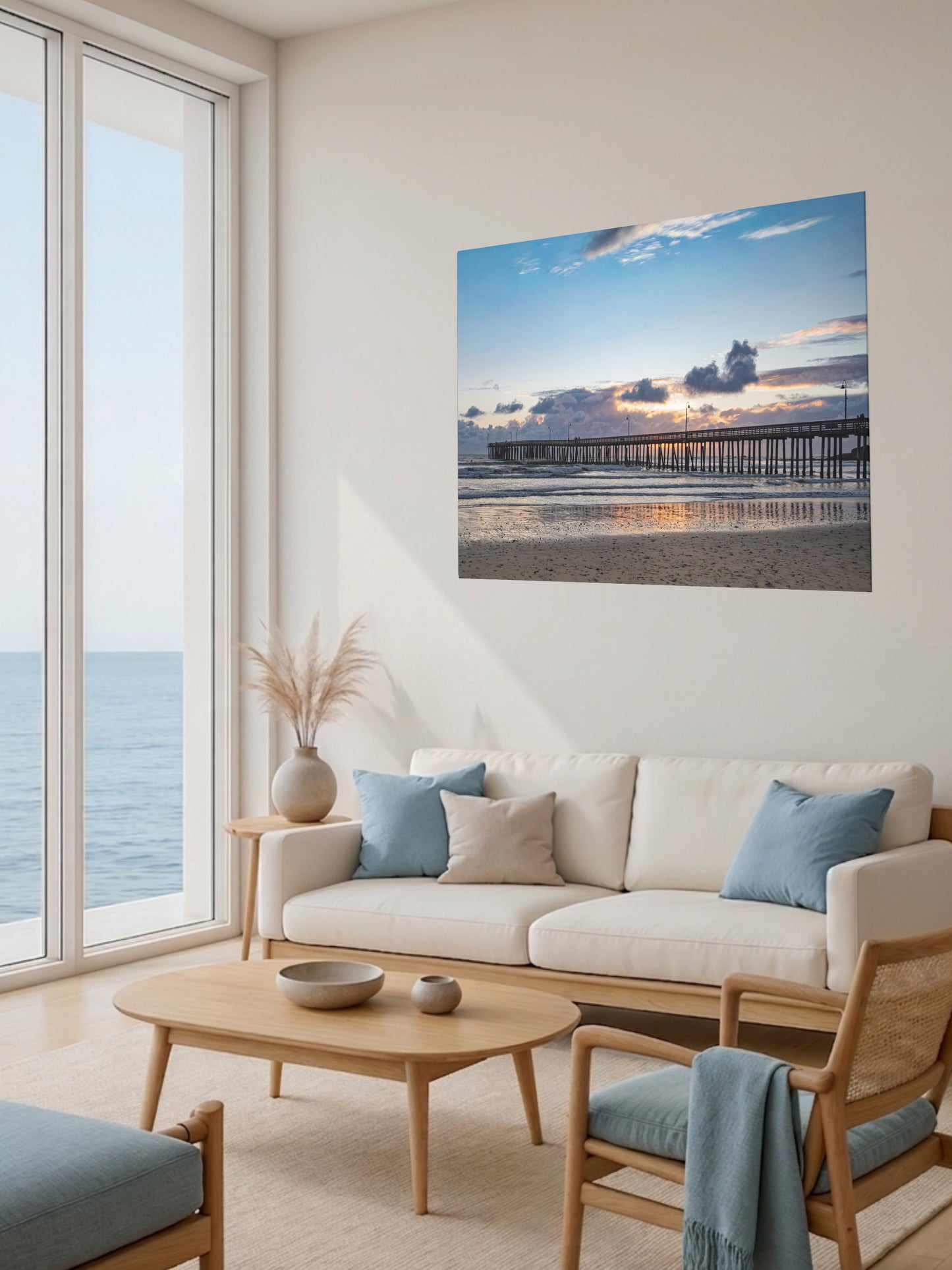 Modern living room with a large window view of the ocean, a beige sofa with blue cushions, and a Cayucos Pier sunset canvas print.