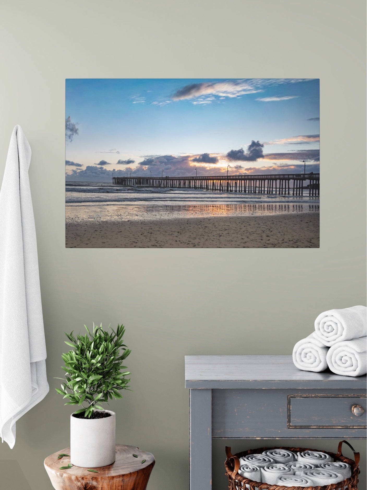 Cayucos Pier sunset canvas print on a wall above a table with a plant and folded towels.