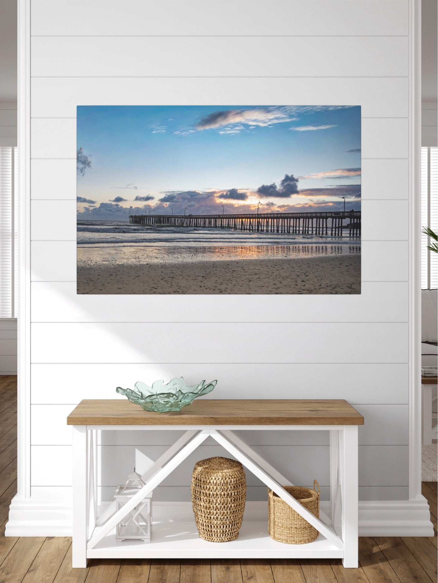 Cayucos Pier sunset canvas print on a wall above a wooden bench with a glass bowl and baskets.
