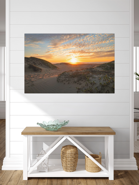 A large canvas print of a beach dunes sunset on a white wall over a wooden bench.