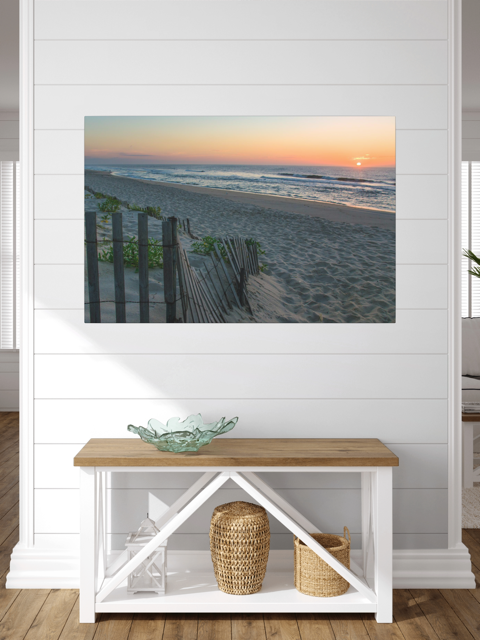 A canvas print of a coastal sunrise on a sandy beach with a weathered wooden fence on a wall above a wooden bench with storage.