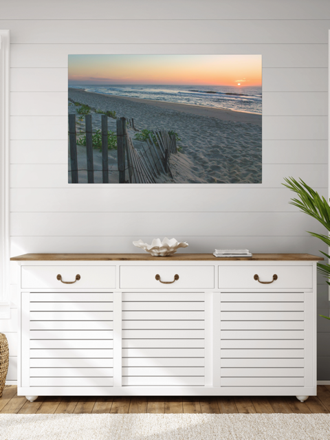 White storage cabinet with wooden top in a room with a canvas print of a coastal sunrise on a sandy beach with a weathered wooden fence on the wall.