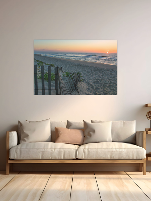 A canvas print of a coastal sunrise on a sandy beach with a weathered wooden fence above a sofa in a room with wooden floor.