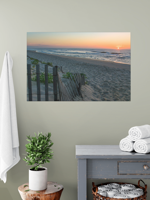 A canvas print of a coastal sunrise on a sandy beach with a weathered wooden fence on a wall above a table with towels and a plant.