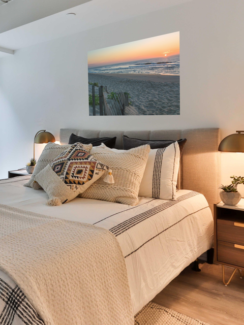 Bedroom with a bed featuring patterned pillows and a canvas print of a coastal sunrise on a sandy beach with a weathered wooden fenceon the wall.