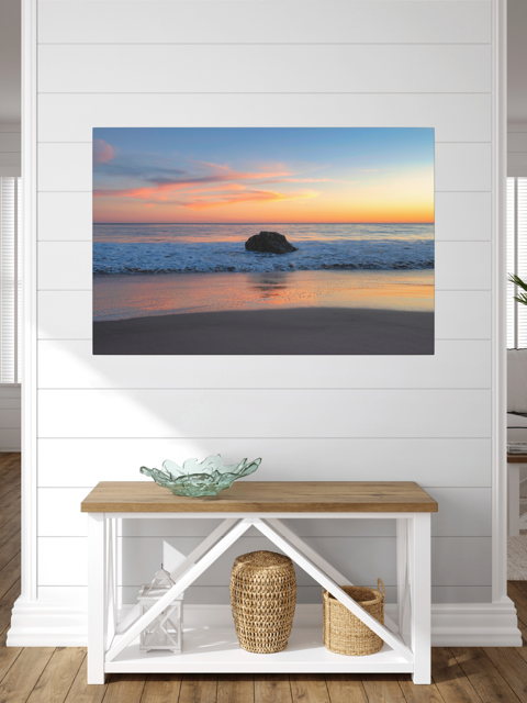 A canvas print of a colorful sunset behind an ocean rock on a wall above a wooden bench with storage.