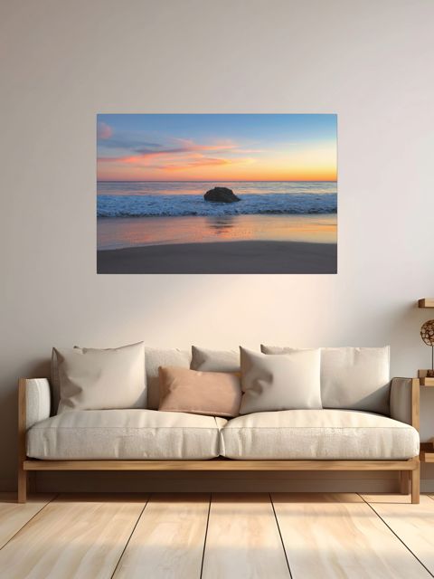 A canvas print of a colorful sunset behind an ocean rock above a sofa in a room with wooden flooring.