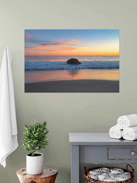 A canvas print of a colorful sunset behind an ocean rock on a wall above a table with a plant and folded towels.