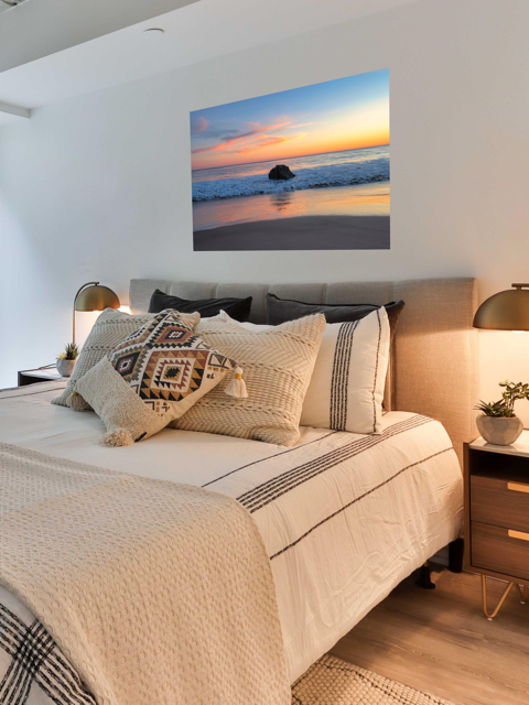 Bedroom with a large bed and decorative pillows, featuring a canvas print of a colorful sunset behind an ocean rock on the wall.