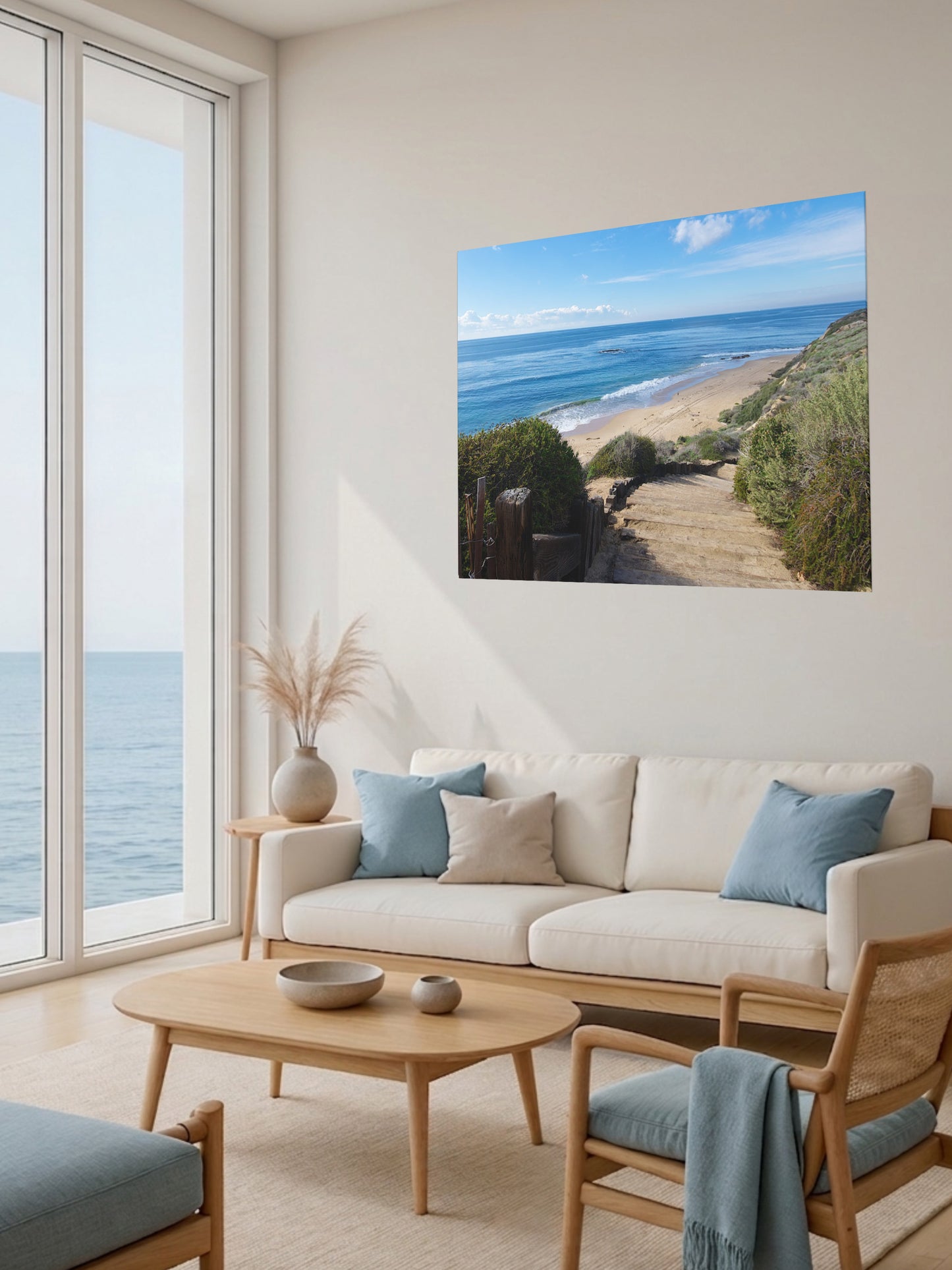 Living room with a large window view of the ocean, a beige sofa with blue cushions, and a canvas print of the wooden stairs leading to Crystal Cove State Beach in Orange County California.