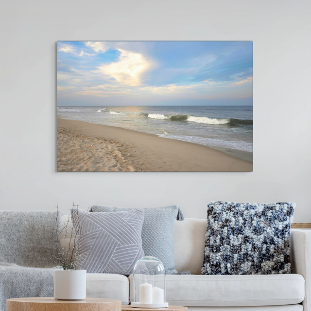 Canvas print of a wide-angle view of the Jersey Shore beach at early evening with rolling ocean waves and a bright, cloudy sky on a wall above a sofa with decorative pillows.