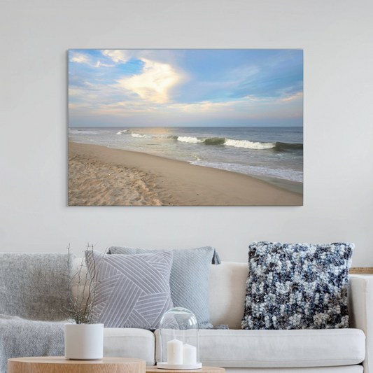 Canvas print of a wide-angle view of the Jersey Shore beach at early evening with rolling ocean waves and a bright, cloudy sky on a wall above a sofa with decorative pillows.