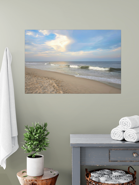 Canvas print of a wide-angle view of the Jersey Shore beach at early evening with rolling ocean waves and a bright, cloudy sky on a wall above a table with a plant and folded towels.