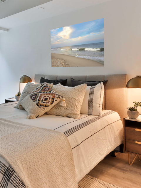 Bedroom with a bed featuring decorative pillows and a canvas print of a wide-angle view of the Jersey Shore beach at early evening with rolling ocean waves and a bright, cloudy sky.