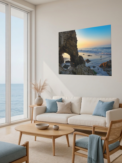 Living room with a large window view of the ocean, a beige sofa with blue cushions, and a canvas print of a sea cave at sunrise in Malibu.