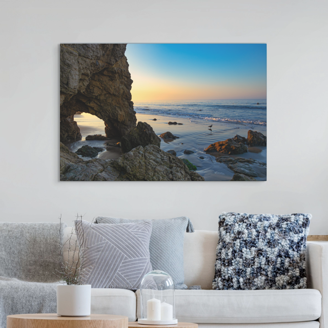 Canvas print of a sea cave at sunrise in Malibu on a wall above a sofa with decorative pillows.