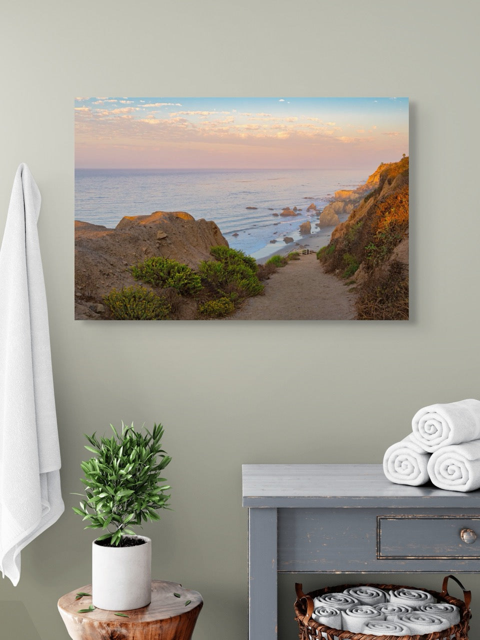 Canvas print of a coastal landscape hanging on a wall above a side table with towels and a plant.
