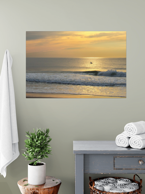 Canvas print of a bird in flight over the ocean at sunriseon a wall above a table with a plant and folded towels.