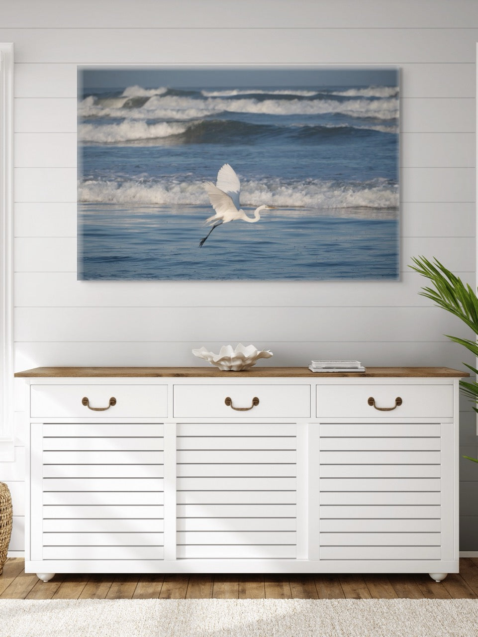 White cabinet with wooden top below a large canvas print of a white egret in flight over the ocean on a white wall.