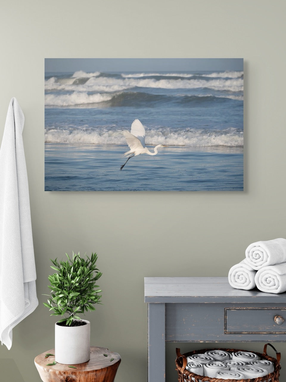 Canvas print of a white egret in flight over the ocean hanging on a wall above a side table with towels and a plant.