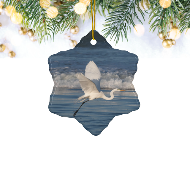 Ceramic Christmas ornament featuring a photograph of a white egret in flight over blue ocean waves with misty sea spray, hanging from a tree.