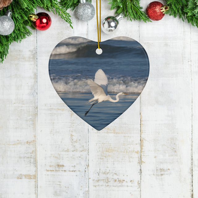 Heart-shaped ceramic Christmas ornament featuring a photograph of a white egret in flight over blue ocean waves with misty sea spray, hanging on a rustic wooden surface with Christmas decorations.