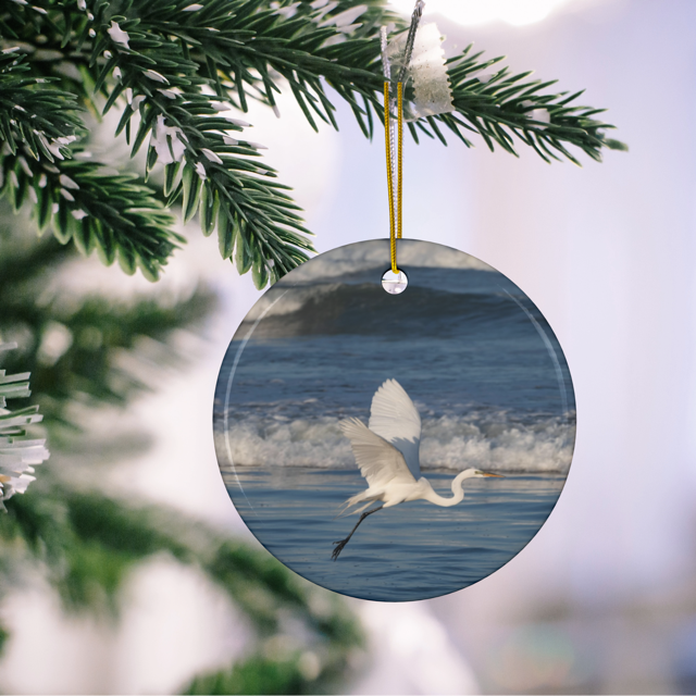 Ceramic Christmas ornament featuring a photograph of a white egret in flight over blue ocean waves with misty sea spray hanging on a tree branch.