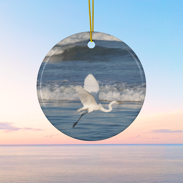 Ceramic Christmas ornament featuring a photograph of a white egret in flight over blue ocean waves with misty sea spray on a blue background