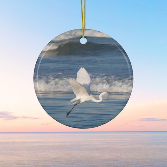 Ceramic Christmas ornament featuring a photograph of a white egret in flight over blue ocean waves with misty sea spray on a blue background
