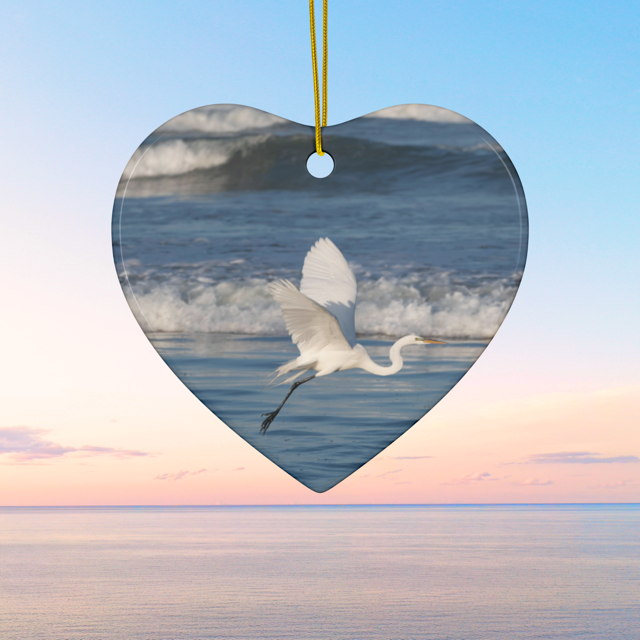 Heart-shaped ceramic Christmas ornament featuring a photograph of a white egret in flight over blue ocean waves with misty sea spray against a blue sky.
