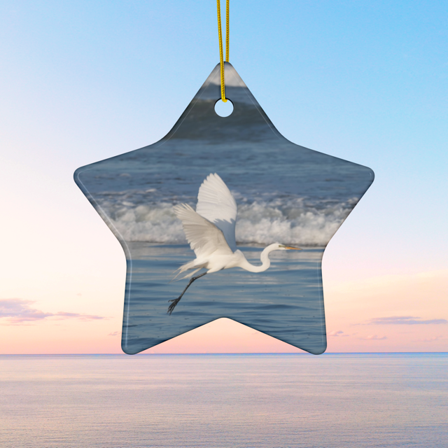 Star-shaped ceramic Christmas ornament featuring a photograph of a white egret in flight over blue ocean waves with misty sea spray. against a sunset sky.