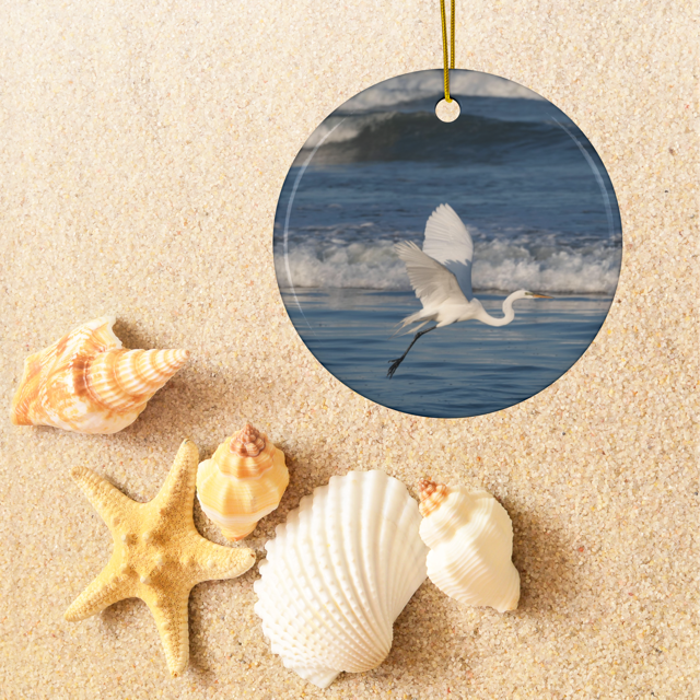 Ceramic Christmas ornament featuring a photograph of a white egret in flight over blue ocean waves with misty sea spray, surrounded by seashells on sand.