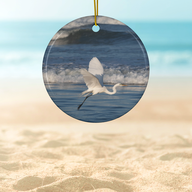 Ceramic Christmas ornament featuring a photograph of a white egret in flight over blue ocean waves with misty sea spray, hanging against a beach background.