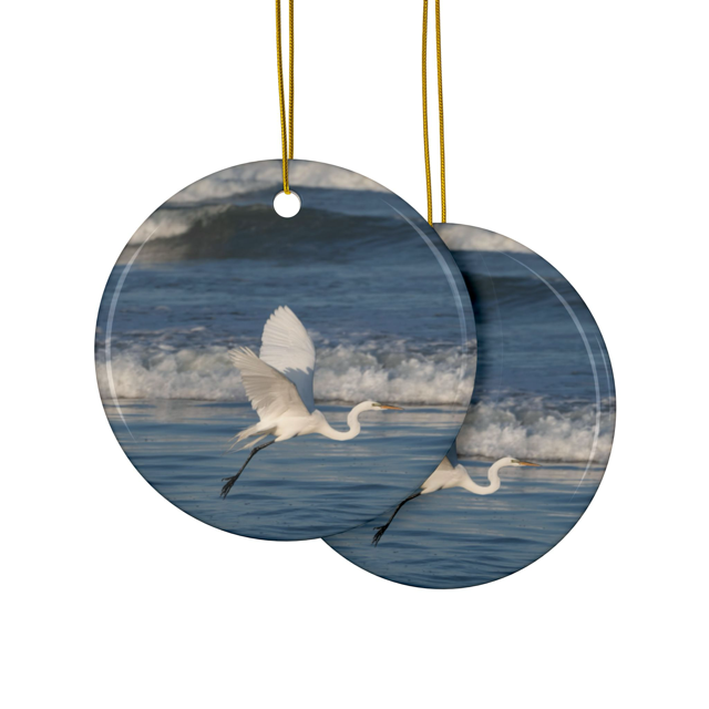 Front and back side of a ceramic Christmas ornament featuring a photograph of a white egret in flight over blue ocean waves with misty sea spray