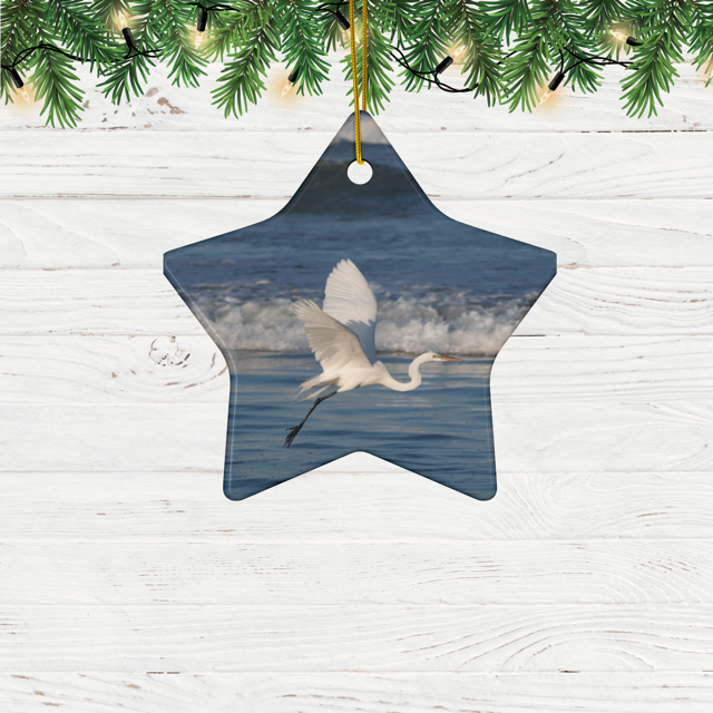 Star-shaped ceramic Christmas ornament featuring a photograph of a white egret in flight over blue ocean waves with misty sea spray. hanging from a Christmas tree.