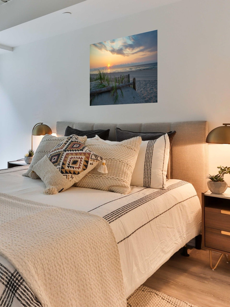 Bedroom with a large bed and decorative pillows, featuring a canvas print featuring a sunrise over the ocean as seen from behind the dunes on the Jersey Shore on the wall.