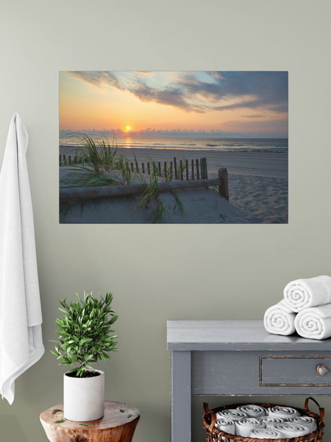 A canvas print featuring a sunrise over the ocean as seen from behind the dunes on the Jersey Shore above a dresser with towels and a plant.