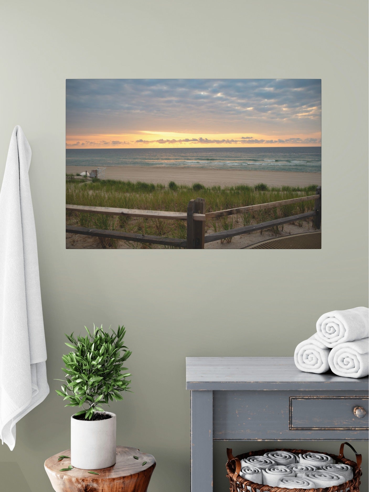 Canvas print of a Jersey Shore sunrise over the Atlantic Ocean with beach dunes and grass on a wall above a dresser with towels and a plant.