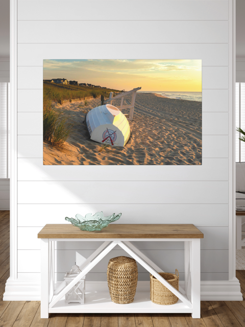 Canvas print of an overturned Jersey Shore Beach Patrol boat and lifeguard stand on the sand at sunrise with beach grass and rolling dunes, above a wooden bench and decorative items in a room.